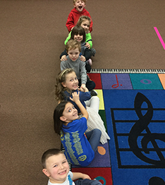 Kids sit forming a line on a carpet with decorative musical notes